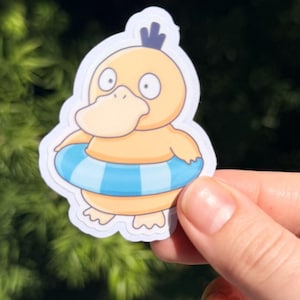 May include: A die-cut sticker featuring a cartoon Psyduck character with a yellow body, a blue and white striped inner tube, and a blue head crest. The sticker is held against a blurred green background.