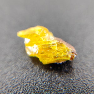 May include: A small, rough-cut mineral specimen with a vibrant yellow hue. The crystalline structure is visible, with a mix of golden and amber tones. The edges have a brownish tint, and the overall shape is irregular.