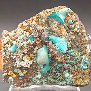 Op de afbeelding: Een mineraal specimen met een ruw, getextureerd oppervlak. De steen toont een verscheidenheid aan kleuren, waaronder donkerbruin, wit, geel en groen. Levendige turquoise formaties zijn aanwezig, wat een opvallend contrast creëert.