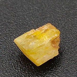 May include: A small, translucent yellow crystal with a rough, uneven surface. The crystal has a rectangular shape with a slightly angled edge. A small, brown area is visible on one side. The crystal is set against a dark, textured background.