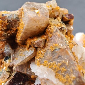 May include: Close-up of a mineral specimen featuring clusters of translucent, light brown crystals. The crystals are accented with golden-orange and white mineral deposits, creating a textured, natural appearance. The overall composition showcases geological formations.