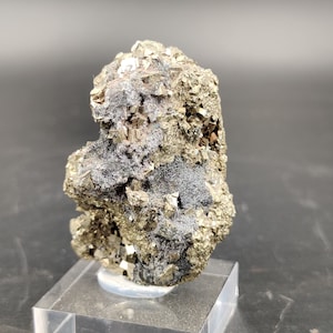Può includere: Un esemplare di pirite naturale, nota anche come oro degli stolti, con un colore oro metallico e una superficie ruvida e strutturata. La pirite è parzialmente ricoperta da un materiale fibroso grigio scuro. L'esemplare è montato su una base acrilica trasparente.
