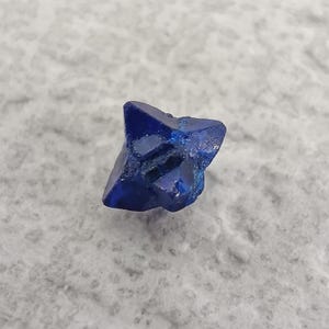 May include: A deep blue, star-shaped crystal with sharp edges. The gemstone has a faceted appearance, reflecting light. The crystal is a rich, saturated blue color, and appears to be a natural mineral specimen.