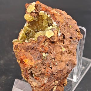 May include: A raw mineral specimen featuring a rough, dark reddish-brown iron-rich matrix with a prominent vug or cavity filled with clusters of small, translucent, pale yellow-green crystals that exhibit a sparkling, crystalline texture.