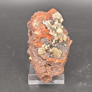 May include: A raw mineral specimen featuring a dark, porous, reddish-brown base with patches of deep charcoal grey. Clusters of small, pale, translucent, crystalline formations are scattered across the upper surface of the rock.