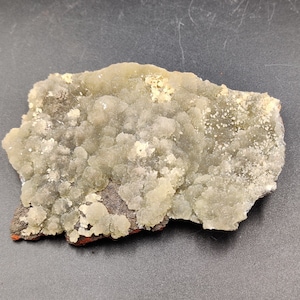 May include: A natural mineral specimen featuring a light green, bubbly surface texture. The mineral has a dark brown base with hints of red and white crystalline deposits, creating a unique geological display.