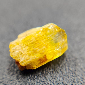 May include: A close-up of a rough, yellow crystal with a translucent appearance. The crystal has a textured surface and a slightly irregular shape, with a mix of bright and muted yellow tones. The background is a blurred, dark gray.
