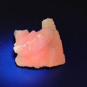 Può includere: Primo piano di un gruppo di formazioni cristalline color pesca. I cristalli hanno una struttura a strati e a blocchi, su uno sfondo blu scuro. I cristalli hanno una consistenza sottile e scintillante.