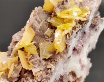 Puede incluir: Primer plano de un espécimen mineral con cristales amarillos vibrantes sobre una matriz de tonos marrón, blanco y rosa. Los cristales tienen una apariencia translúcida. La composición general muestra formaciones geológicas naturales.