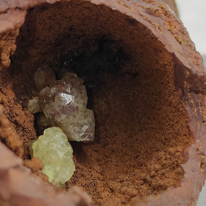 May include: Close-up of a geode with a rough, reddish-brown exterior. The interior reveals clusters of pale green and clear crystals. The crystals have a faceted appearance, suggesting a mineral specimen.