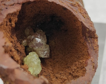 May include: Close-up of a geode with a rough, reddish-brown exterior. The interior reveals clusters of pale green and clear crystals. The crystals have a faceted appearance, suggesting a mineral specimen.