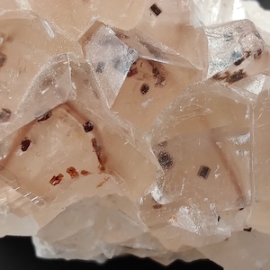 Peut inclure: Gros plan sur un amas de cristaux de calcite translucides de couleur pêche. Les cristaux ont une structure géométrique et carrée avec de petites inclusions brun foncé dispersées. La texture générale est rugueuse et cristalline.