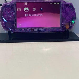 May include: A transparent purple Sony PSP handheld game console on a black stand. The screen displays a pink and purple interface with game and saved data options. The console is in good condition.