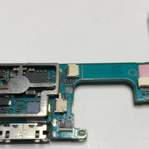May include: A close-up of a green circuit board with various electronic components, including a chip, a connector, and a metal shield. The board has a rectangular shape and is divided into two sections.