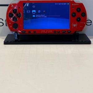 May include: A red Sony PSP handheld gaming console on a black stand. The screen displays a blue interface with icons. The console has a directional pad, buttons, and a spider graphic. The PSP is a portable entertainment device.