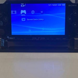 May include: A black Sony PlayStation Portable handheld gaming console featuring a bright blue screen display with menu icons, the text Saved Data Utility, and a green power indicator light, mounted on a black rectangular display stand.