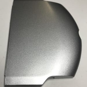 May include: Silver-colored plastic cover for a handheld gaming device. The cover has a smooth, slightly textured surface.