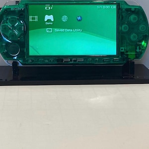 May include: A transparent green Sony PSP handheld game console on a black stand. The screen displays a green interface with icons for Game and Saved Data Utility. The console has a glossy finish and visible buttons.