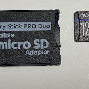 May include: Black memory stick pro duo adapter with the text "Memory Stick PRO Duo compatible micro SD Adaptor". A 128GB microSD card with the text "TEAMGROUP Team 128 GO micro A1 U3 V30 1" is shown next to the adapter.