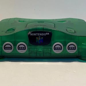 May include: A transparent green Nintendo 64 game console. The console has a curved design with rounded edges and features the Nintendo 64 logo. The front has four controller ports and the text "NINTENDO 64".