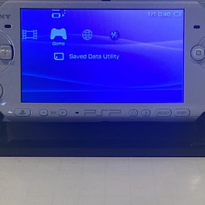 May include: A silver Sony PSP handheld game console on a black stand. The screen displays a blue interface with game and saved data utility icons. The console has a directional pad, buttons, and a power indicator.