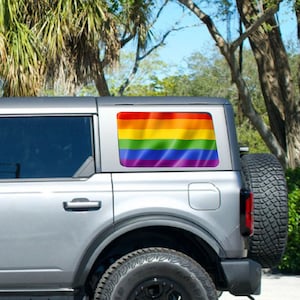 May include: A gray Ford Bronco with a rainbow flag decal on the rear window. The vehicle is parked on a paved surface with palm trees in the background.