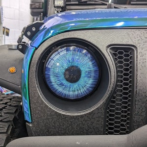 May include: A close-up of a black Jeep Wrangler with a custom blue and green paint job. The Jeep has a black front bumper and a black grille with a honeycomb pattern. The headlights are black and have a blue eye design. The windshield has a sticker that says "DIAMOND VINYL CO. YOURMOMSFAVORITEWRAP"