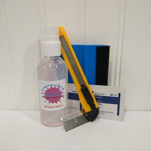 Pu&ograve; includere: Un set di strumenti per applicare adesivi in ​​vinile, tra cui una bottiglia di fluido per l'applicazione del vinile, un coltello multiuso giallo, una spatola blu e una lama da rasoio. La bottiglia ha un'etichetta rosa con il testo "Diamond Vinyl Decal Splash Vinyl Application Fluid Shake Well".