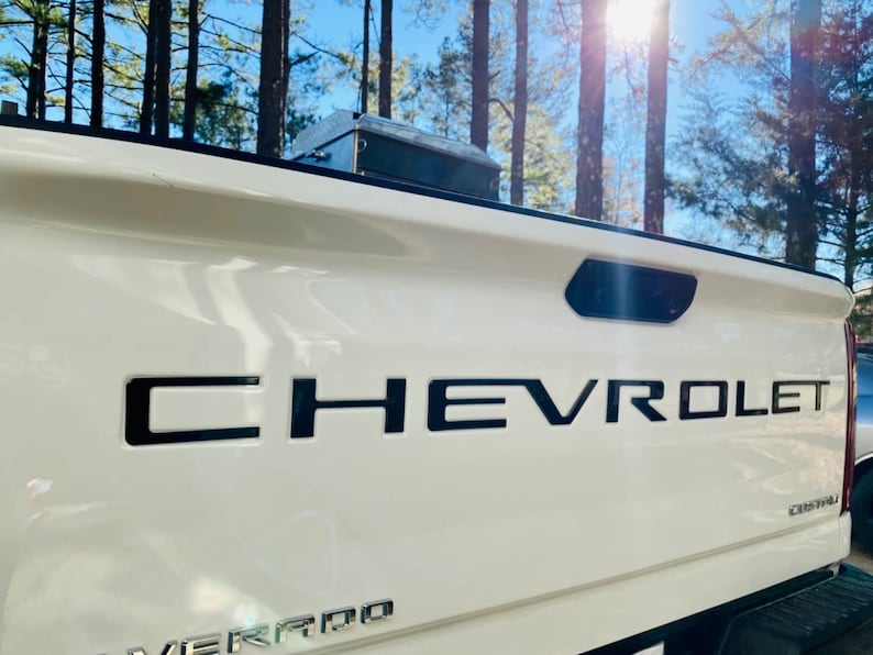 May include: White pickup truck tailgate with a black Chevrolet emblem. The truck has a black rear window and a black taillight.