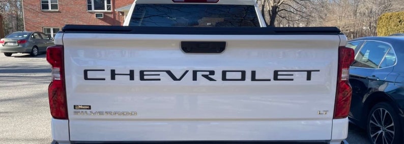 May include: White Chevrolet Silverado pickup truck with a black tailgate and the word "CHEVROLET" in black lettering across the tailgate.