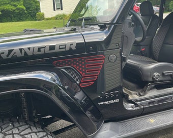 Może przedstawiać: Czarny Jeep Wrangler Unlimited z naklejką amerykańskiej flagi w kolorze czerwonym, białym i niebieskim na błotniku. Pojazd ma czarne koła i czarne wnętrze.