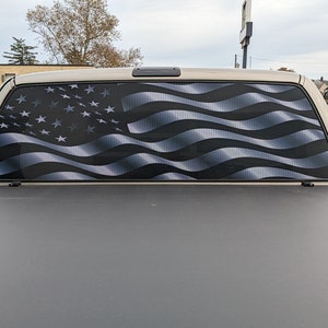 May include: A black pickup truck with a rear window decal of an American flag with a wavy design. The flag is silver and black.
