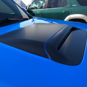 May include: A blue car with a black hood scoop. The scoop has a textured, black, rectangular design.