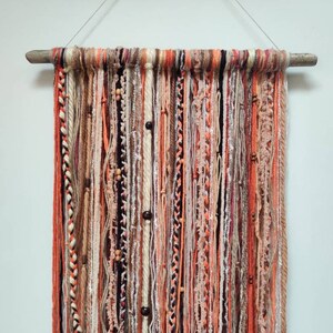 May include: A wall hanging made of yarn in shades of orange, brown, and white. The yarn is woven together to create a textured and layered effect. The hanging is adorned with wooden beads.