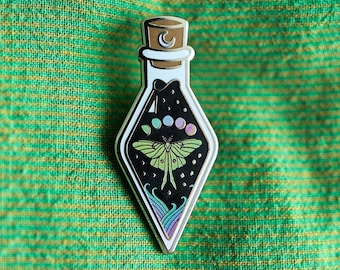 May include: A silver enamel pin shaped like a glass bottle with a cork stopper. The bottle contains a black background with a green and white moth, a crescent moon, and stars.