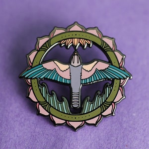 May include: A green, pink, and blue enamel pin featuring a stylized bird with outstretched wings flying inside a circular mandala design.
