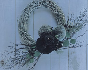 May include: A gray twig wreath with black velvet and lace flowers and a gray velvet pumpkin. The wreath is decorated with green leaves and branches.