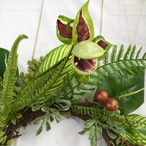May include: A green and brown artificial plant wreath with a large, green, open-mouthed, carnivorous plant in the center. The wreath is made of various types of greenery, including ferns and leaves.
