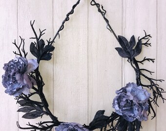 May include: A black twig wreath with three purple velvet flowers. The wreath is hanging on a white wooden door.