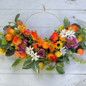 May include: A white metal wreath form decorated with a variety of artificial flowers and fruit. The flowers are in shades of orange, yellow, pink, and white. The fruit is yellow and orange.