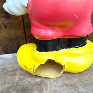 May include: A vintage plastic Mickey Mouse figurine with a yellow shoe that is cracked and broken. The figurine is wearing a red shirt and black belt.