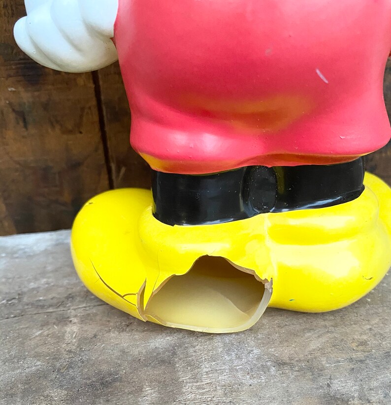 May include: A vintage plastic Mickey Mouse figurine with a yellow shoe that is cracked and broken. The figurine is wearing a red shirt and black belt.