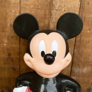 May include: A black and red plastic Mickey Mouse figurine. The figurine is standing with its arms crossed and has a big smile on its face.