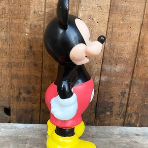 May include: A black and red plastic Mickey Mouse figurine with yellow shoes. The figurine is standing on a wooden surface.