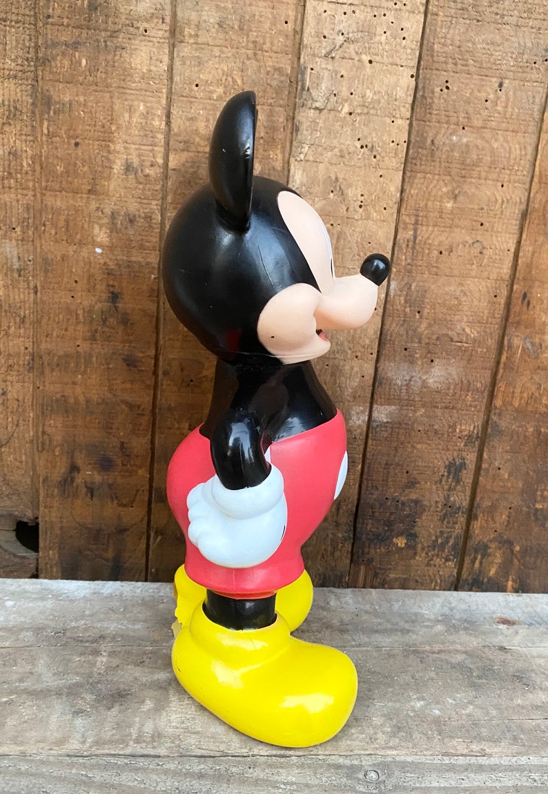 May include: A black and red plastic Mickey Mouse figurine with yellow shoes. The figurine is standing on a wooden surface.