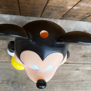 May include: Black and white plastic Mickey Mouse figurine with a yellow base. The figurine has a hole in the top of its head.