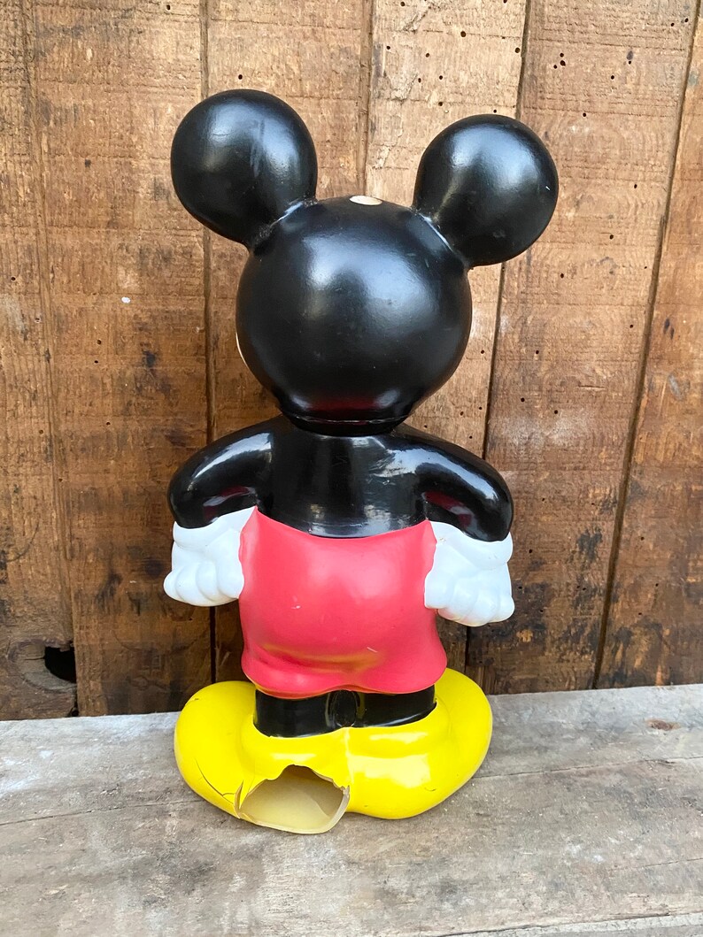 May include: A vintage black and red Mickey Mouse figurine with yellow shoes. The figurine is made of plastic and has a chipped shoe.
