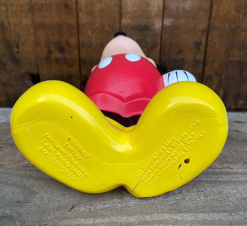 May include: Yellow and red plastic Mickey Mouse bath toy. The toy has a red shirt with black outlines and two white circles. The toy's feet are yellow and have the following text: "DISNEY MPG by Monogram Products Inc. Largo, Florida 34649 MADE IN CHINA NOT DISHWASHER SAFE WASH THOROUGHLY BEFORE USING WASH IN WARM WATER ONLY AVOID TEMPERATURE EXTREMES"
