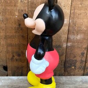 May include: A black and red plastic Mickey Mouse figurine with yellow shoes. The figurine is standing on a wooden surface.