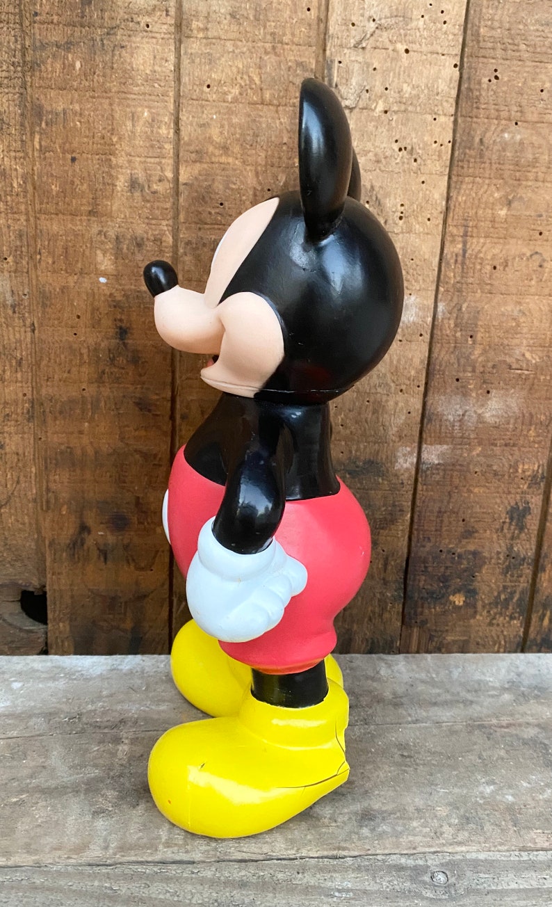 May include: A black and red plastic Mickey Mouse figurine with yellow shoes. The figurine is standing on a wooden surface.